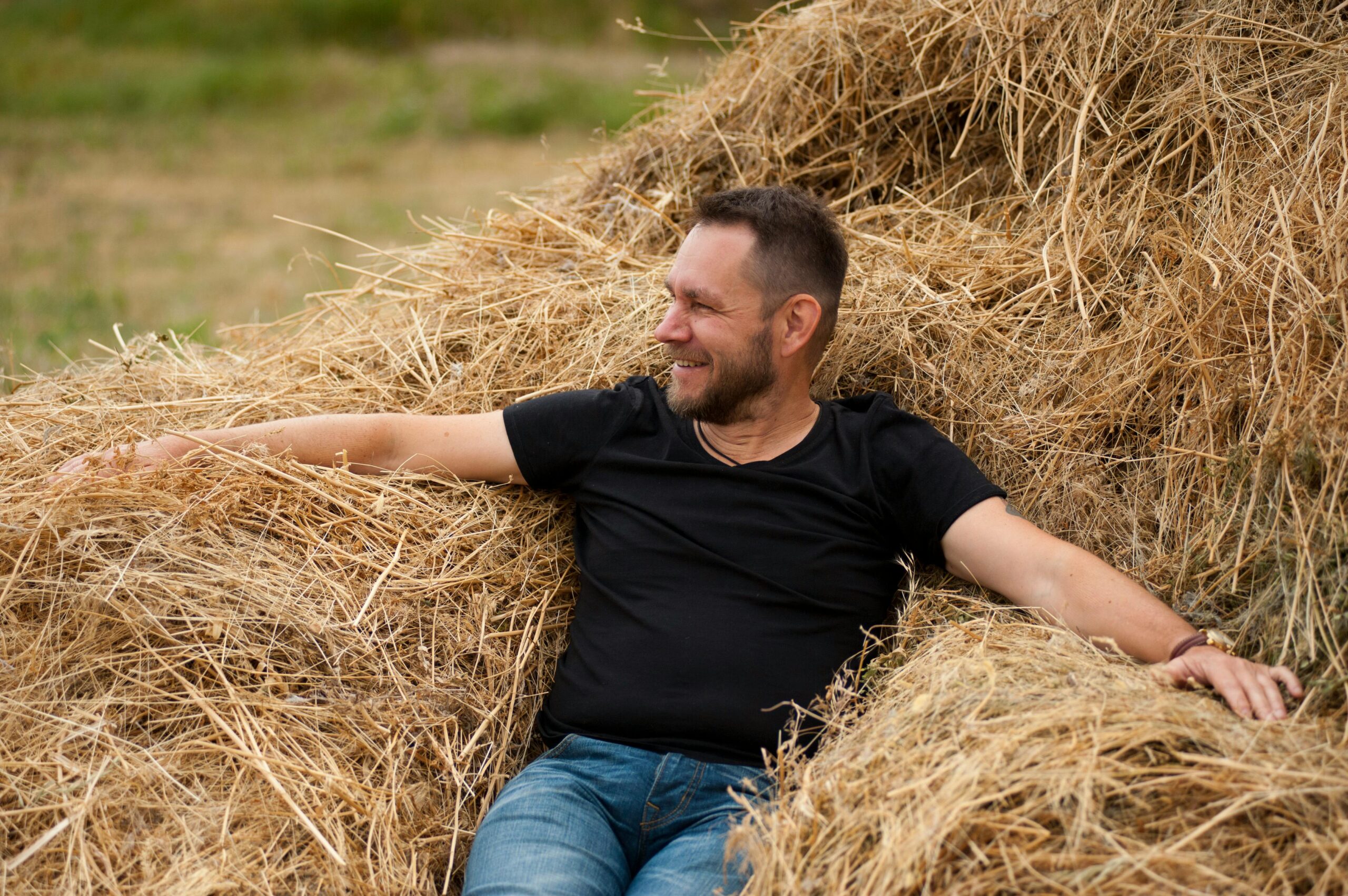 A man in a black t-shirt smiles while relaxing in a haystack amidst a peaceful countryside setting.